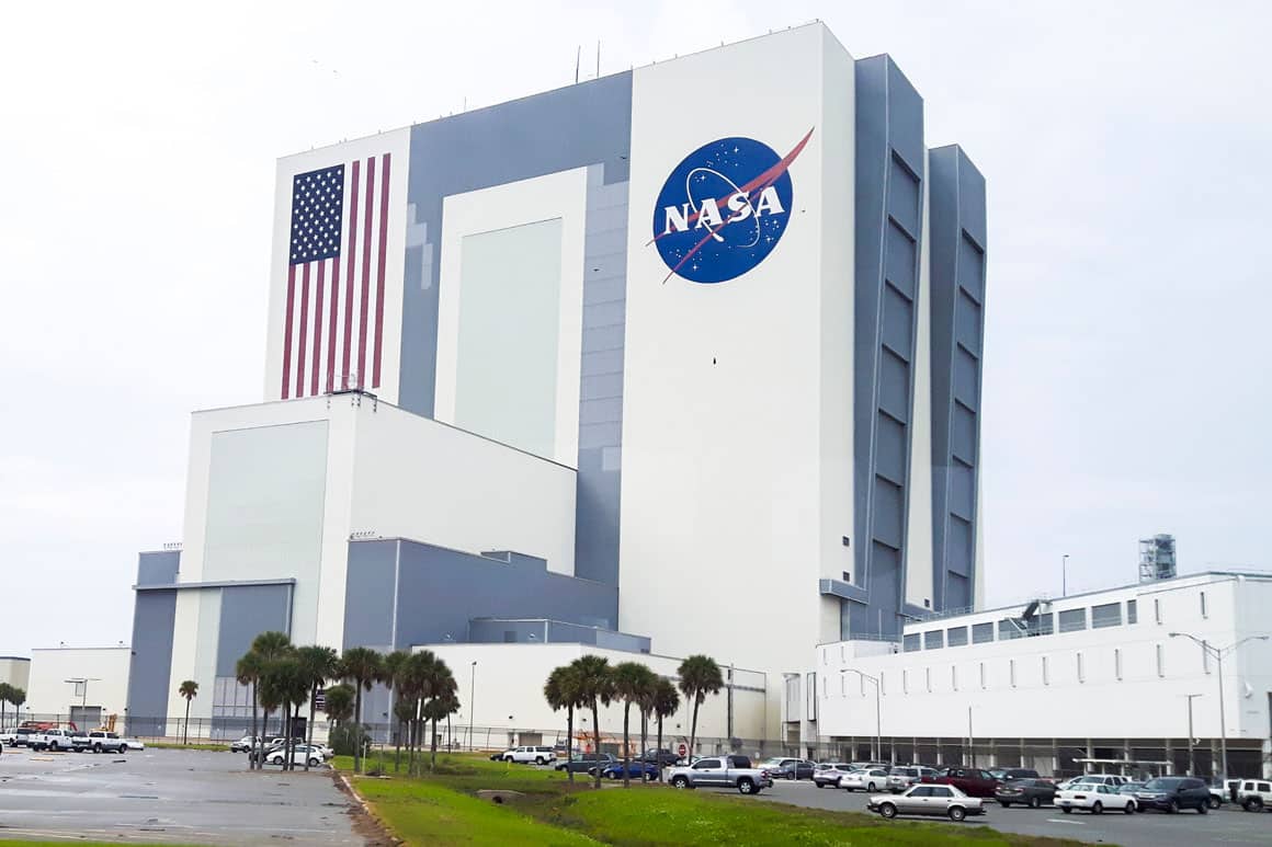 Vista ampla das instalações do Kennedy Space Center na Flórida: prédios industriais da NASA, Vehicle Assembly Building ao fundo, áreas de lançamento e paisagem da Costa Espacial sob céu nublado.