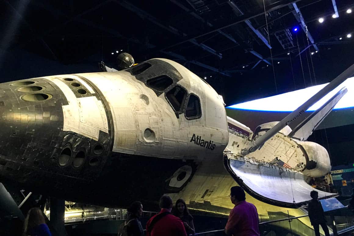 Ônibus espacial Atlantis original em exposição no Kennedy Space Center, suspenso com portas de carga abertas e braço robótico estendido, simulando órbita espacial
