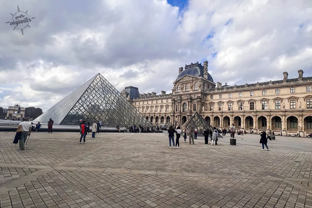 Pirâmide de vidro I.M. Pei no pátio do Museu do Louvre Paris: fachada clássica ao fundo, visitantes e céu parcialmente nublado.