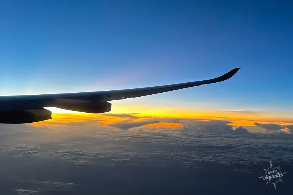Avião comercial em voo sobre nuvens ao pôr do sol: vista das asas e céu laranja durante viagem transatlântica Europa