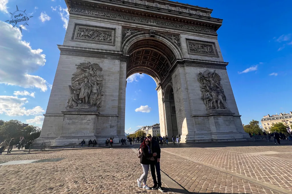 Arco do Triunfo Paris Champs-Élysées: fachada neoclássica com baixos-relevos esculturas Vitória e pedestres casal selfie em praça pavimentada céu azul
