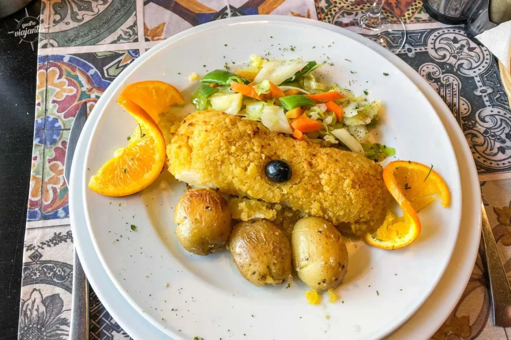 Peixe empanado dourado com crosta de farofa e azeitona preta no centro, acompanhado de batatas cozidas com casca, legumes cozidos (cenoura, bróceli, vagem) e fatias de laranja fresca decorando o prato branco sobre mesa com azulejos coloridos portugueses. Rabelos Restaurante, Vila Nova de Gaia.
