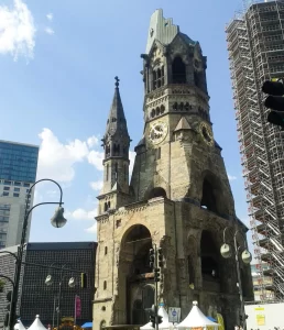 Igreja Quebrada de Berlim durante o dia, torre sineira gótica parcialmente destruída, ao lado da igreja nova com vitrais azuis modernos, ruínas preservadas sob céu claro.