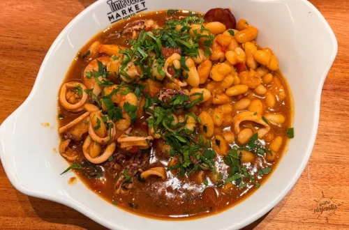 Restaurantes no Porto e Gaia - Feijoada de frutos do mar servida no Restaurante Meia-Nau do Time Out Market em Porto, com feijão branco cremoso, lulas e gambas frescas, azeitonas pretas, coentro abundante e caldo saboroso, em panela de cobre tradicional sobre prato branco.