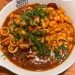 Restaurantes no Porto e Gaia - Feijoada de frutos do mar servida no Restaurante Meia-Nau do Time Out Market em Porto, com feijão branco cremoso, lulas e gambas frescas, azeitonas pretas, coentro abundante e caldo saboroso, em panela de cobre tradicional sobre prato branco.