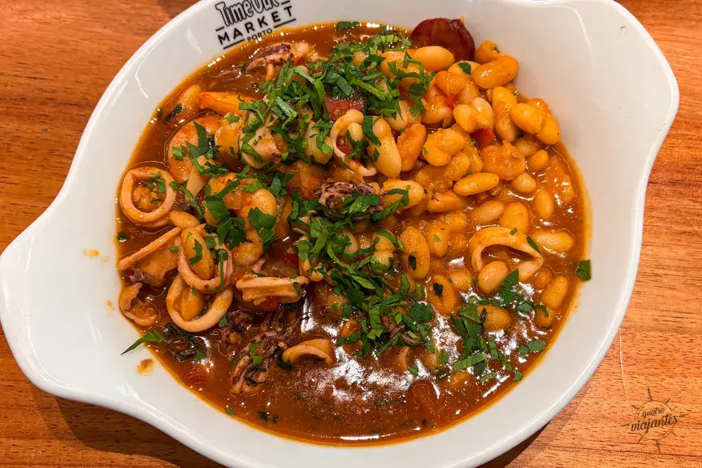 Restaurantes no Porto e Gaia - Feijoada de frutos do mar servida no Restaurante Meia-Nau do Time Out Market em Porto, com feijão branco cremoso, lulas e gambas frescas, azeitonas pretas, coentro abundante e caldo saboroso, em panela de cobre tradicional sobre prato branco.