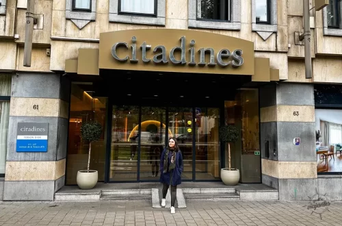 Mulher na entrada Citadines Apart’hotel Toison d'Or Bruxelas, fachada amarela moderna com plantas e portas de vidro.