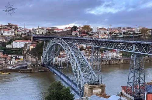 Ponte D. Luís I que liga a cidade do Porto a Vila Nova de Gaia. vista de cima com arco metálico sobre o Rio Douro, edifícios coloridos de Porto e Gaia sob céu parcialmente nublado.