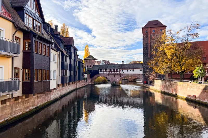 Paisagem da Nuremberg medieval, Alemanha, mostrando a ponte de madeira coberta Henkersteg sobre o rio Pegnitz. À esquerda, edifícios históricos com fachadas de madeira escura refletem-se na água calma. À direita, uma torre de pedra alta e árvores com folhas amarelas de outono sob um céu azul com nuvens esparsas.