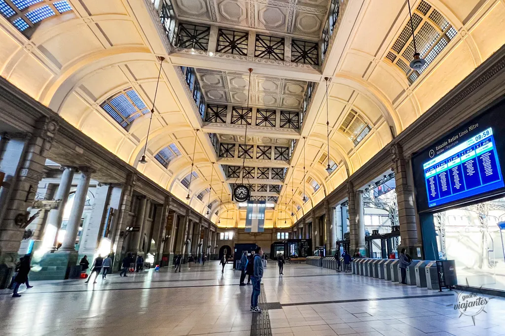 Interior do saguão principal da Estación Retiro - Línea Mitre em Buenos Aires, mostrando o fluxo de passageiros e o painel digital de horários dos trens