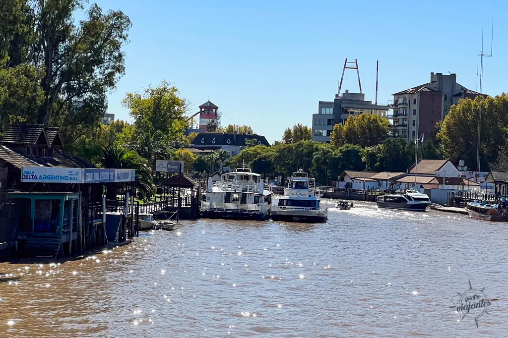 O que fazer Em Tigre? Barcos de passeio e catamarãs ancorados no rio em Tigre, Buenos Aires, com o píer da Líneas Delta Argentino à esquerda sob um céu ensolarado.